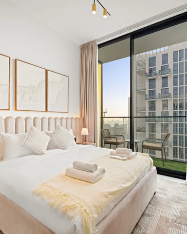 Modern bedroom with cream upholstered headboard, white bedding, and floor-to-ceiling windows overlooking city buildings