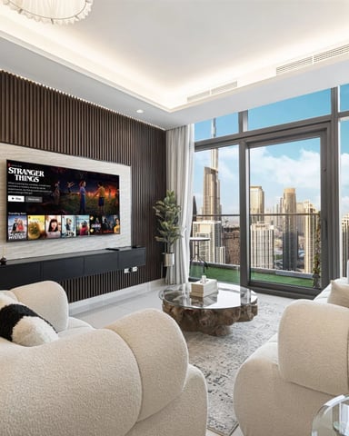Modern living room with cream curved seating, wall-mounted TV, floor-to-ceiling windows overlooking city skyline, and wood-slat accent wall.