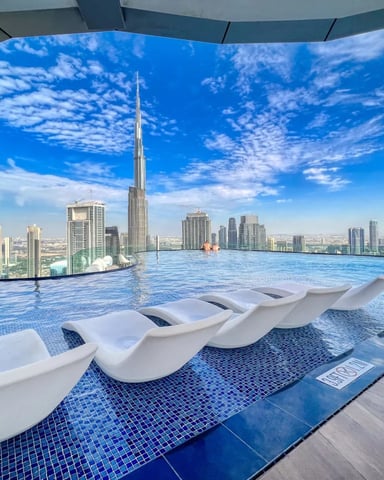 Rooftop infinity pool with white loungers overlooking tall skyscraper and city skyline under blue sky