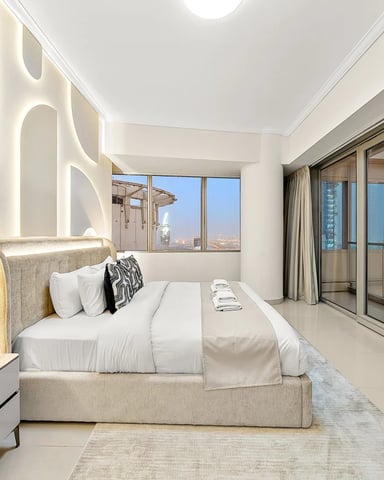 Modern high-rise bedroom with upholstered bed, sculptural wall art, floor-to-ceiling windows overlooking cityscape and distant buildings