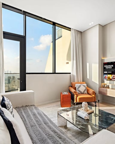 Modern bedroom with floor-to-ceiling windows, white sofa, orange leather chair, wall-mounted TV, and city skyline view