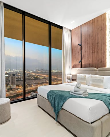 Modern bedroom with queen bed, floor-to-ceiling windows overlooking cityscape at dusk, wood accent walls, and desk with TV.