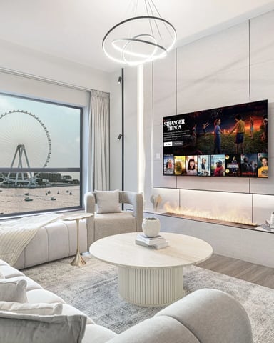 Modern living room with cream furniture, round coffee table, wall-mounted TV, and large window overlooking a ferris wheel and beach