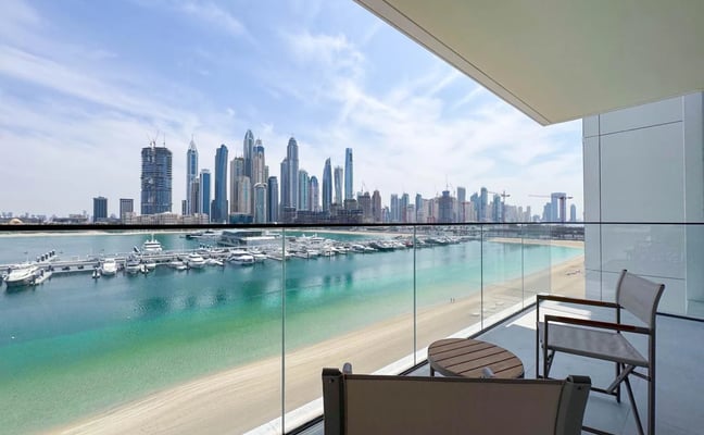 Balcony with lounge chairs overlooking turquoise beach, marina, and high-rise city skyline