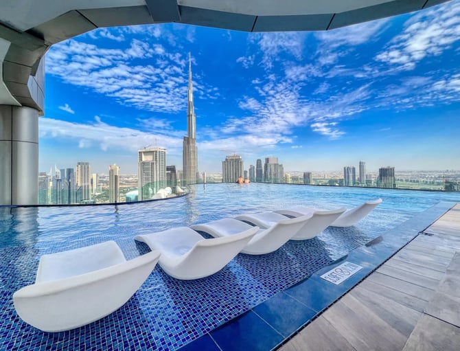 Rooftop infinity pool with white loungers overlooking tall skyscraper and cityscape under blue sky